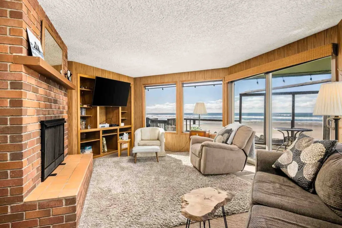 The image showcases a spacious, light-filled living room with a crackling fireplace, plush seating, and floor-to-ceiling windows framing breathtaking ocean views. The warm, inviting atmosphere is perfect for relaxing, socializing, and soaking in the scenic beauty of Rockaway Beach.