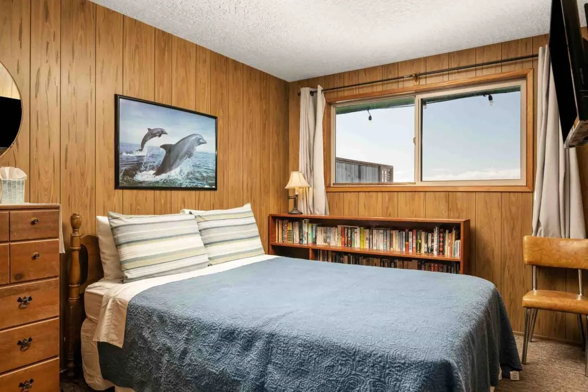 This interior image features a cozy bedroom with a bed covered in a soothing blue coverlet, playful dolphin artwork above the headboard, a well-stocked bookshelf, a soft lamp providing warm lighting, and a window with curtains letting in natural light. The arrangement exudes comfort and a welcoming ambiance, ideal for relaxation and unwinding.