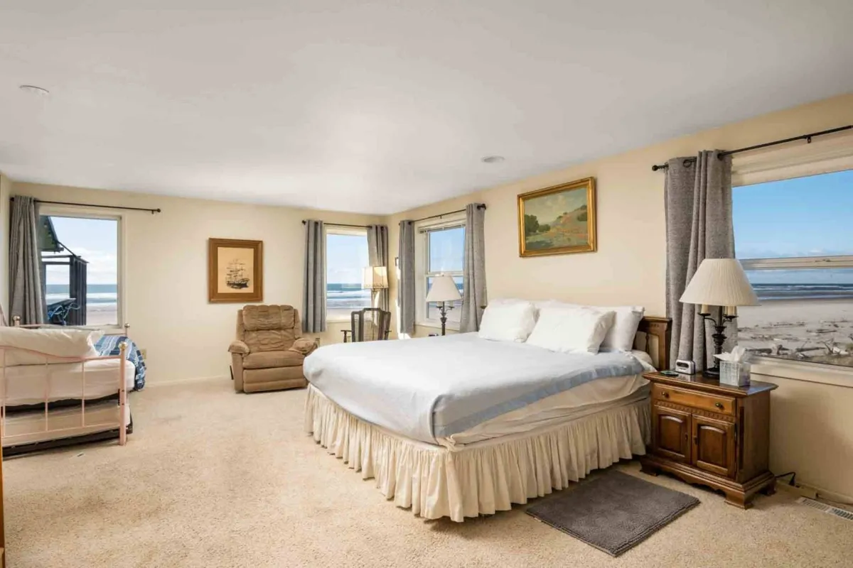 A spacious bedroom with a large bed, windows that offer glimpses of the outdoors, a comfy recliner for reading, and artistic touches throughout. The layout suggests both relaxation and the opportunity to enjoy tranquil views from within.