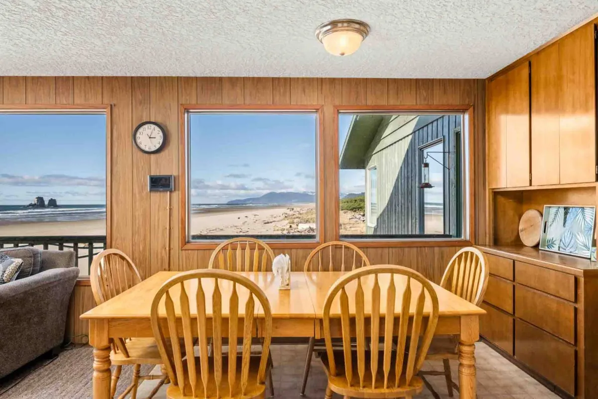 This image captures a cozy dining area featuring a large table perfectly positioned beside expansive windows. Outside, a stunning beach view pours in, filling the room with natural light. Warm wood paneling and a prominent clock add to the inviting, mid-century-inspired ambiance. The space is clearly designed for gathering, sharing meals, and enjoying the ocean vista.