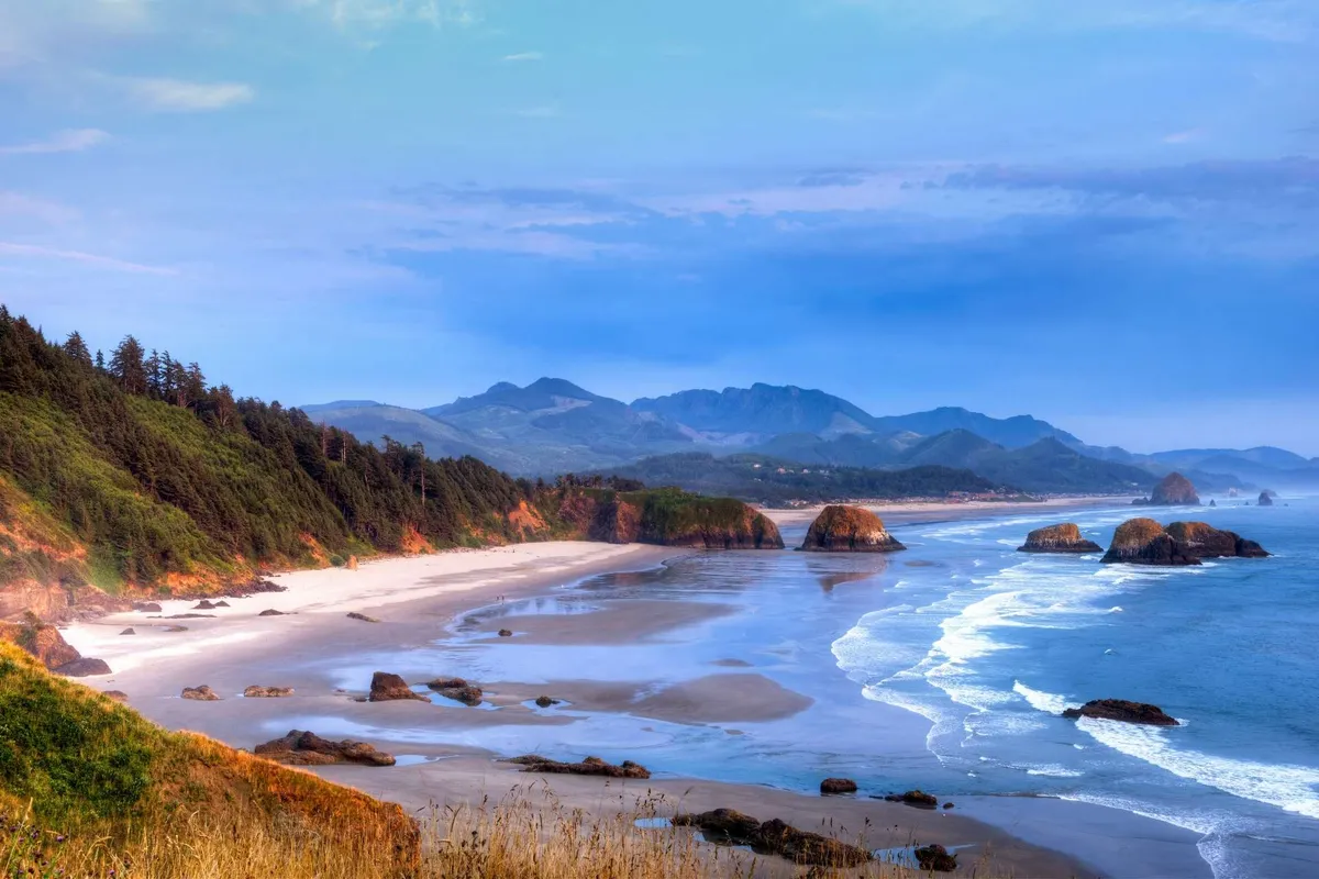 This image showcases a sweeping view of the coastline at sunset, with gentle ocean waves lapping against the sandy beach. Rock formations and mountains rise in the distance, while the forest edges frame the shoreline. People can be seen walking along the water’s edge, and tide pools are visible in the foreground, inviting exploration. The mood is peaceful and family-friendly, perfect for safe outdoor fun.
