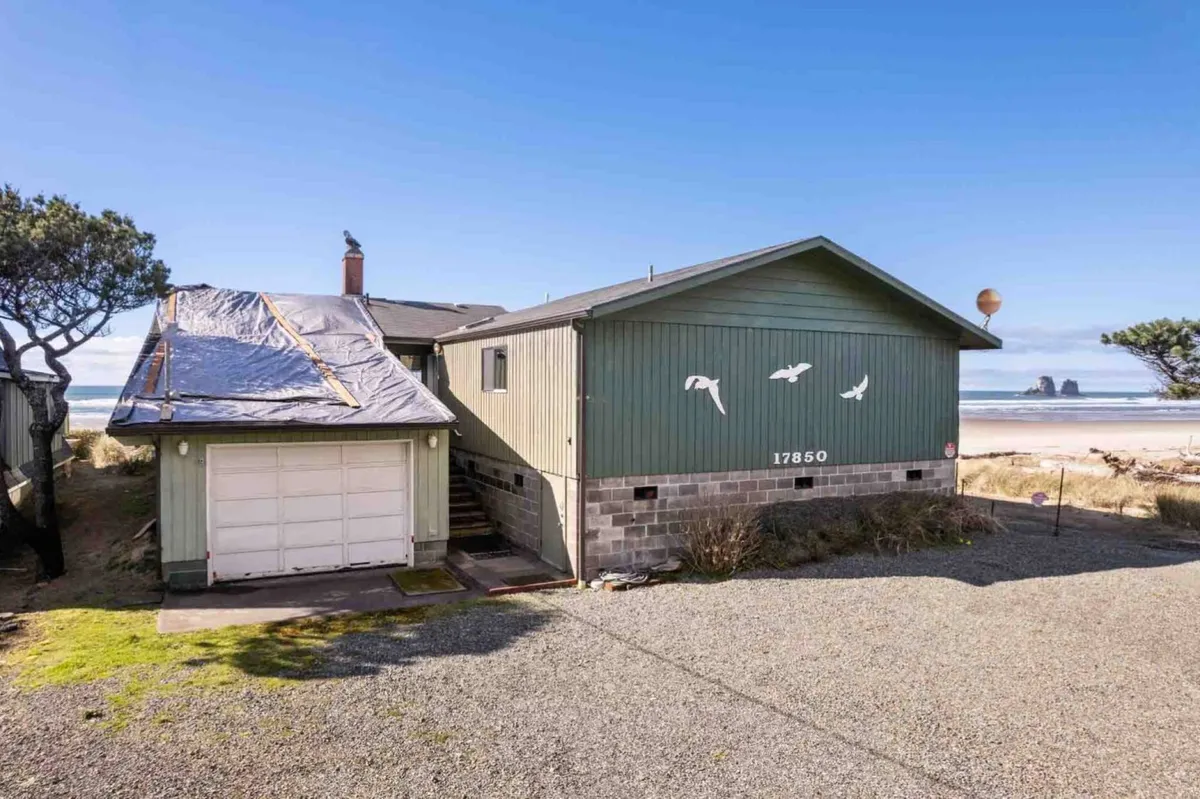 An exterior view highlighting the home's green siding, adjacent garage, and the ocean just beyond, framed by lush coastal trees. The setting exudes a relaxed mood, inviting guests to imagine beach walks or gatherings with the soothing sound of the waves in the background. The horizon hints at early morning or late afternoon light, emphasizing connection to the outdoors.
